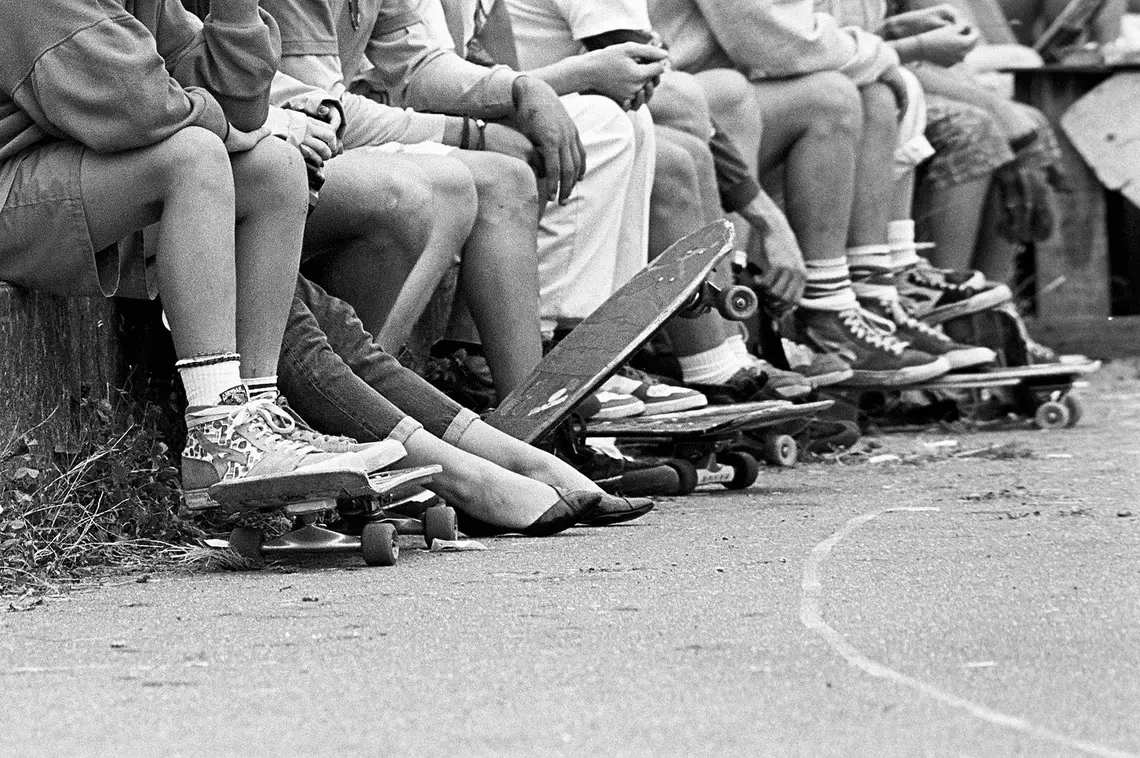 Imagem em preto e branco de várias crianças dos anos 50 sentadas na calçada com alguns skates nos pés.