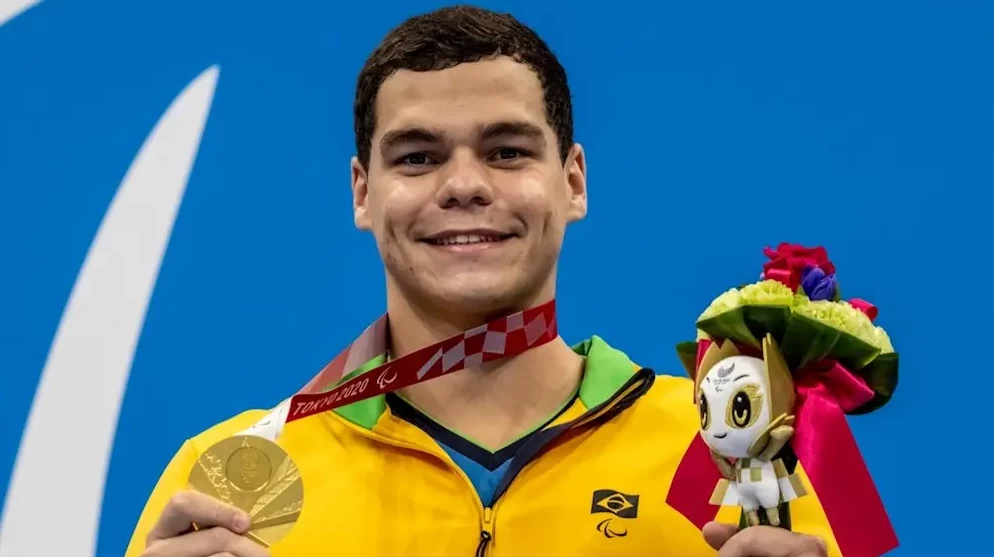 Gabriel Bandeira, um dos maiores atletas paralímpicos do Brasil, vestido com blusa do Brasil, segurando medalha em uma mão e boneco das paralimpíadas