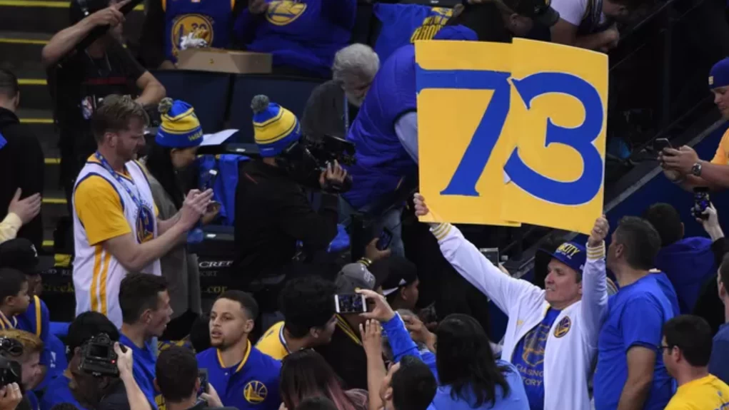 foto tirada da arquibancada em um jogo do Warriors, onde um homem levanta placas com os números 7 e 3, formando "73", que indica um dos recordes da NBA do time