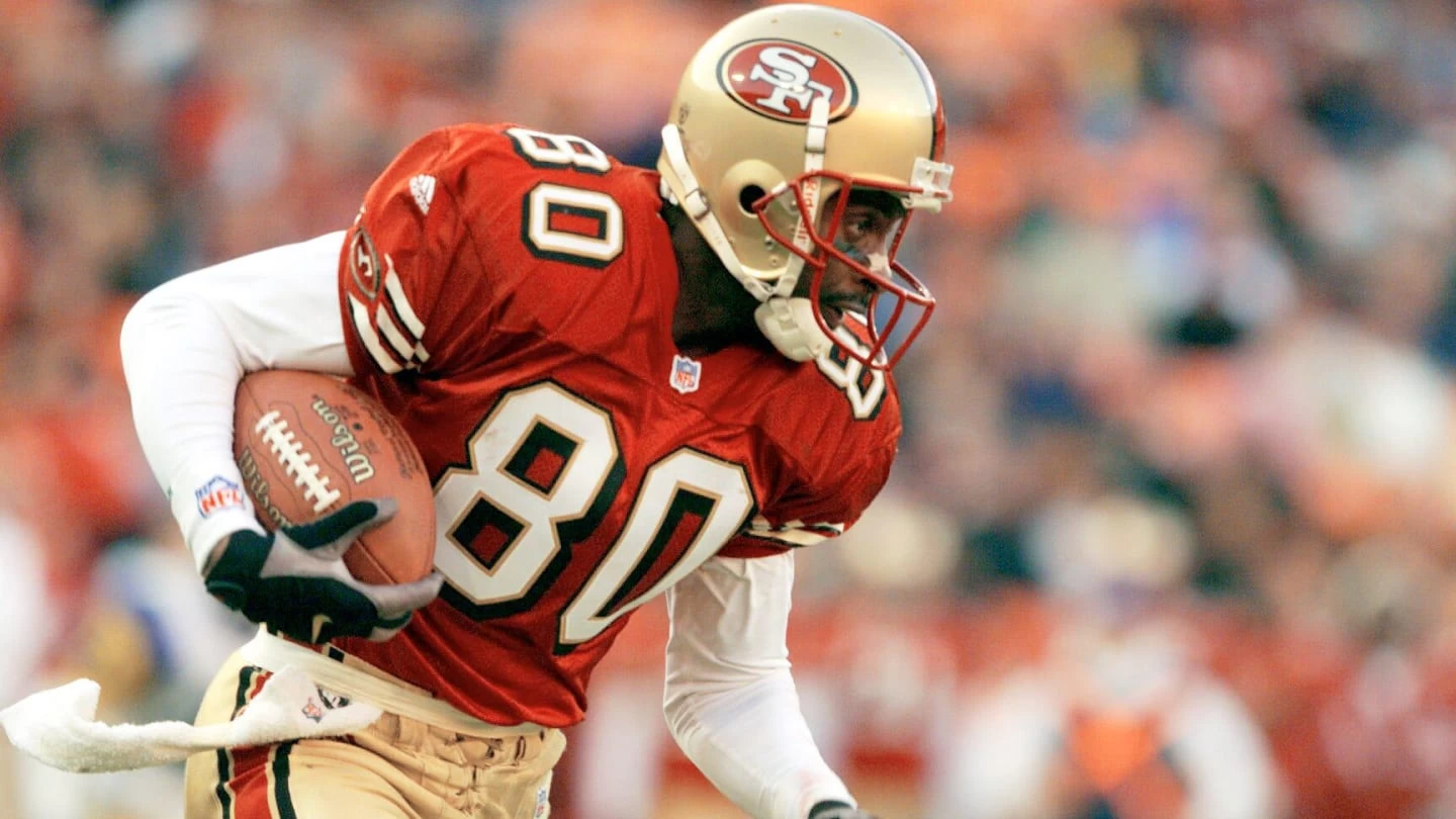 Jerry Rice, uma das lendas da NFL, em posição de ataque, em campo, segurando uma bola de futebol americano